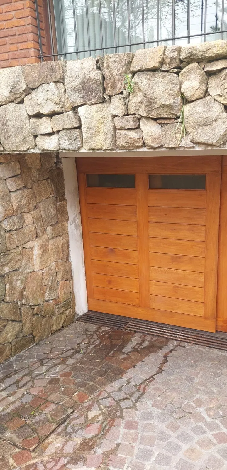 Portón de garaje de dos hojas en madera clara, con visores, bajo un muro de piedra